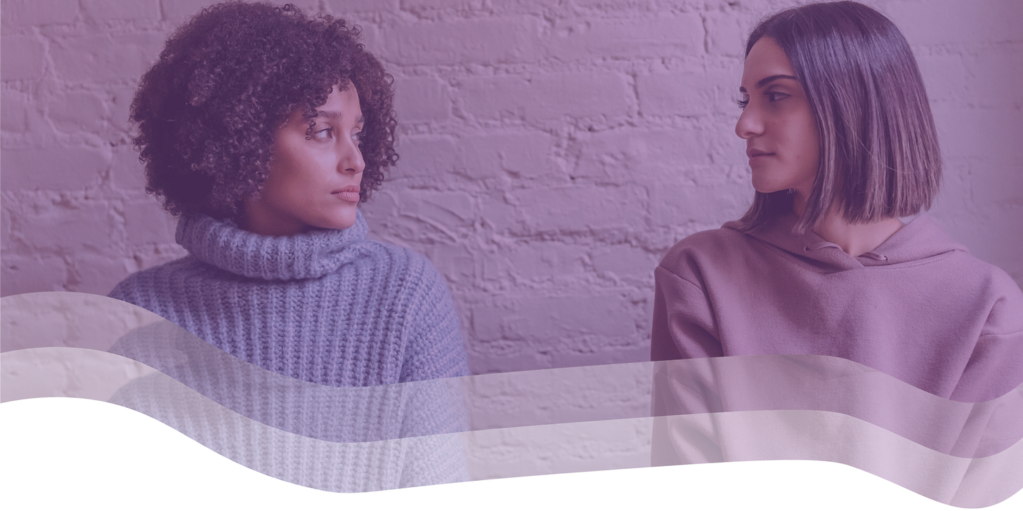 two women look at each other, unsmiling, against a white brick background.