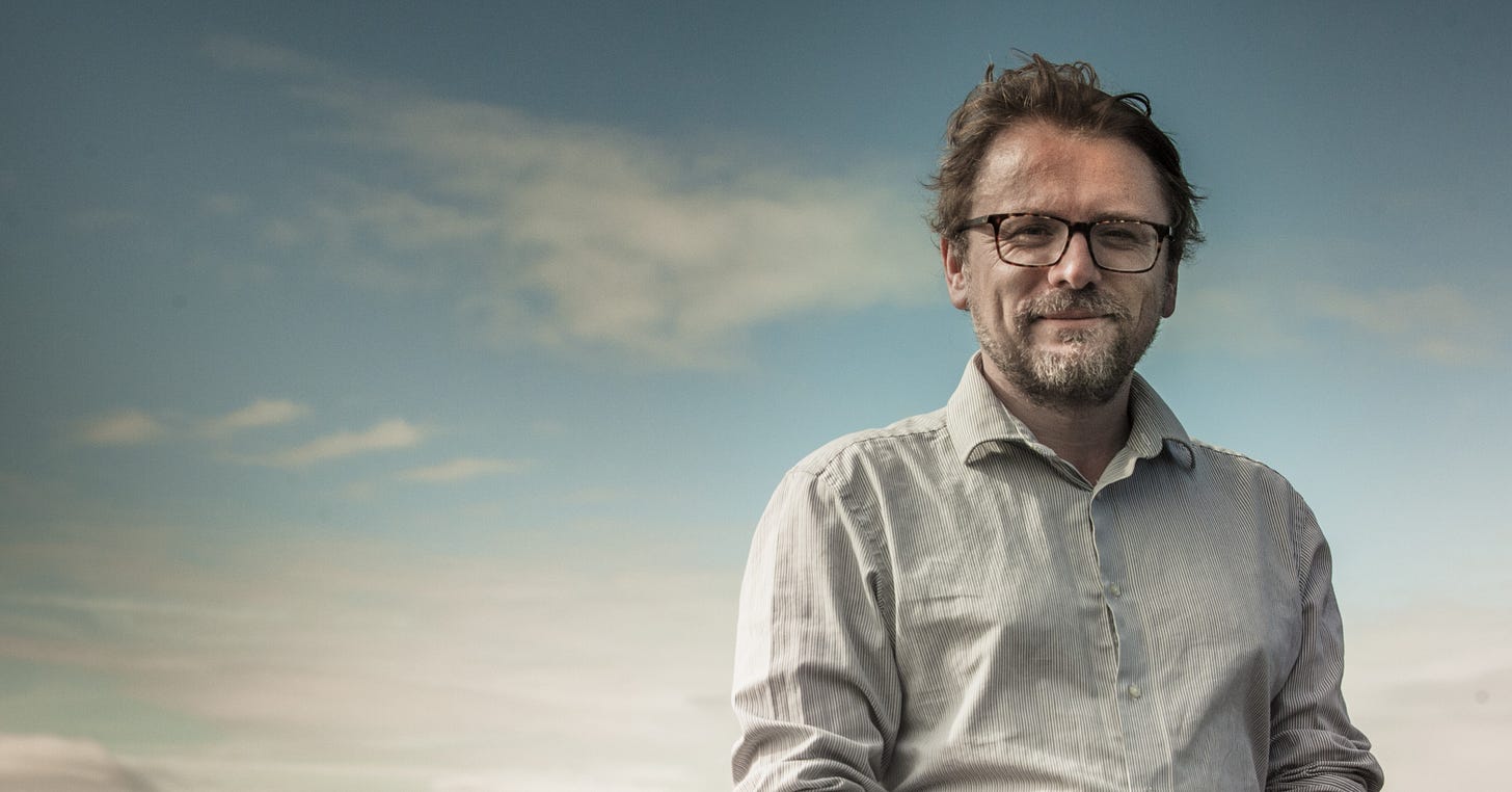 A photograph of Andy Hamilton wearing a lightly coloured shirt and glasses, standing against a blue sky with some clouds A photograph of Andy Hamilton wearing a lightly coloured shirt and glasses, standing against a blue sky with some clouds