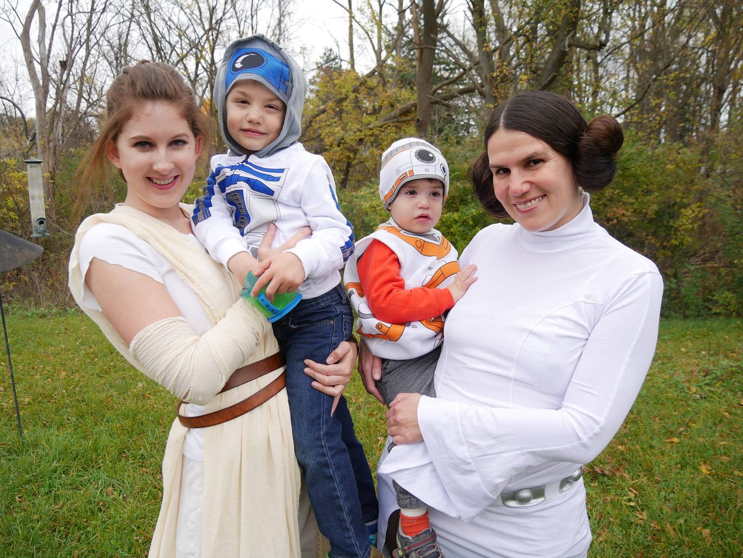 A woman dressed like Rey holds a boy in an R2-D2 sweatshirt. Next to her a woman dressed like Leia holds a toddler dressed like BB-8. It is daytime and they’re outside with fall trees behind them.