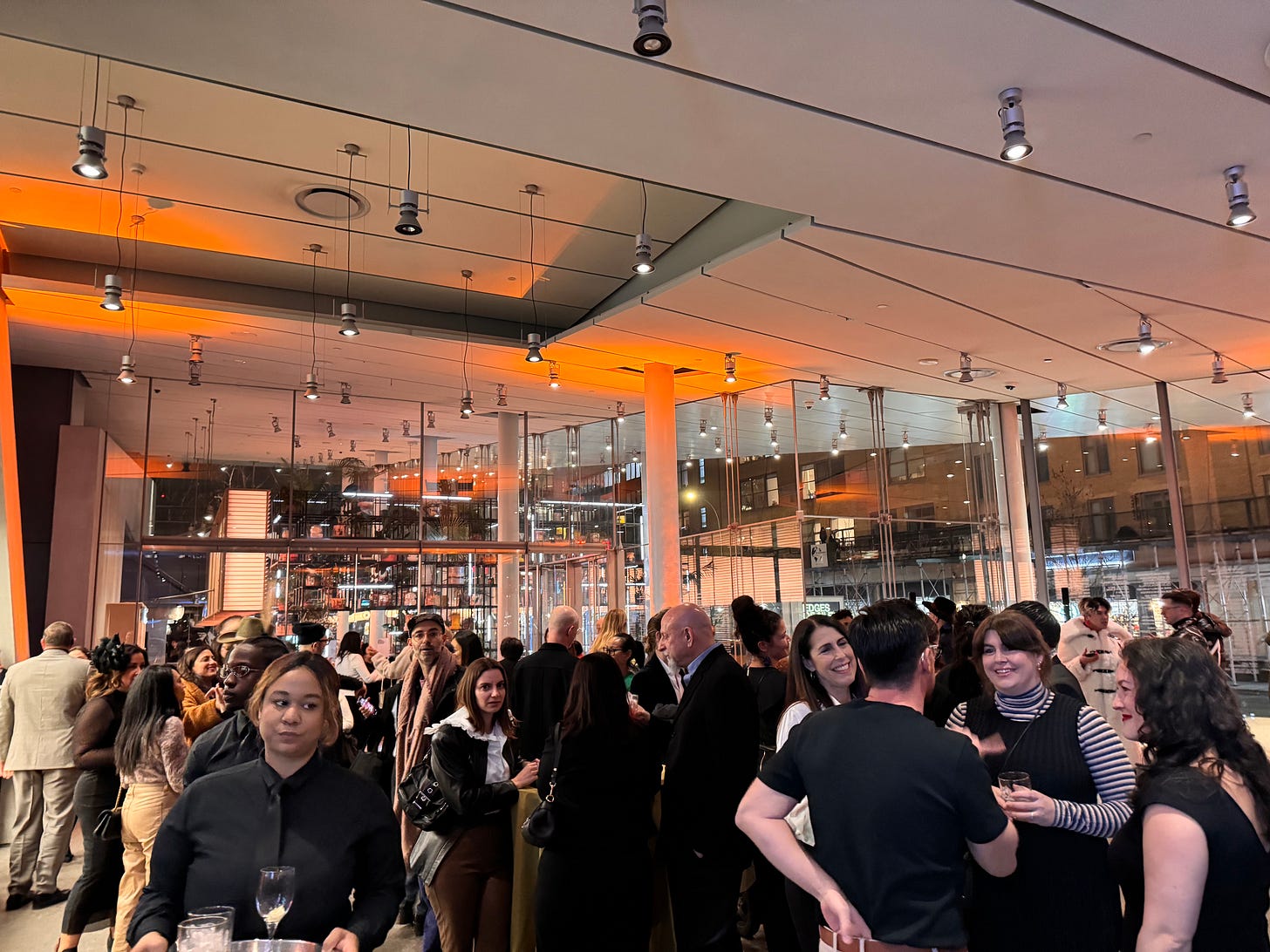The crowd on the first level of the Whitney Museum of American Art. Evening lights are on and people stand chatting over drinks.