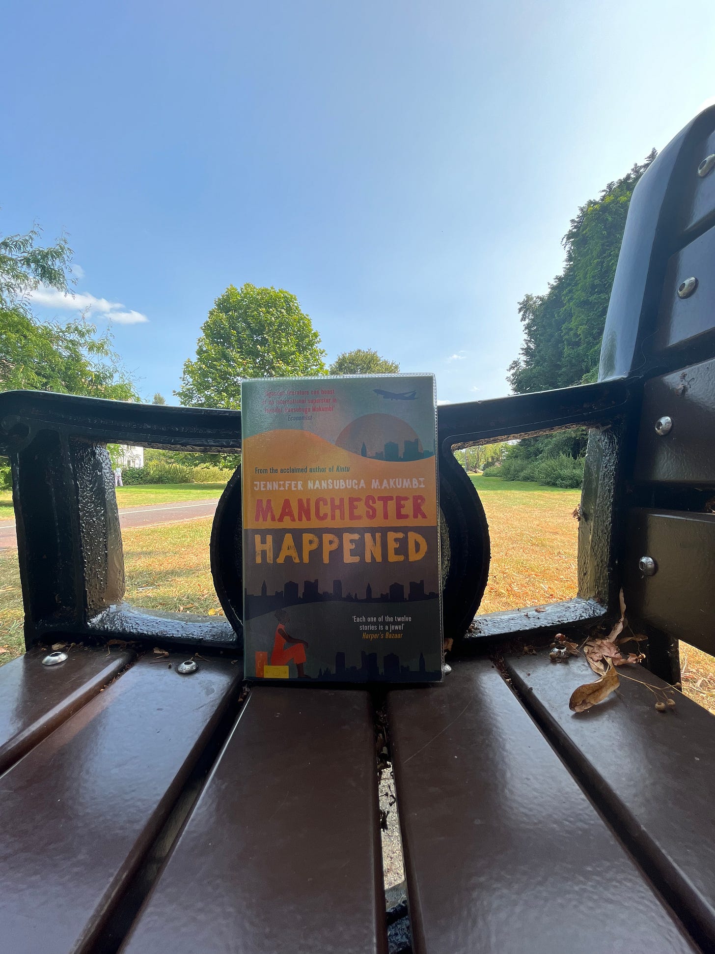 Manchester Happened by Jennifer Nansubuga Makumbi. book cover in green, yellow and red. Background: arm of a bench with a lush green park behind Manchester Happened by Jennifer Nansubuga Makumbi. book cover in green, yellow and red. Background: arm of a bench with a lush green park behind