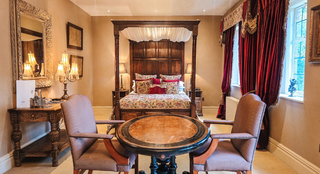 A hotel room with a four-poster bed, a dining table, a large mirror, and a side table with a lamp on top