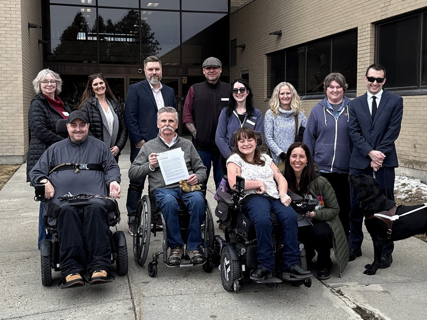 A group of disability advocates in manual wheelchairs, power, wheelchairs, service animal, and non apparent disabilities standing outside the accessible entrance of the montana supreme court. a man in wheelchair is holding up a letter to the attorny general