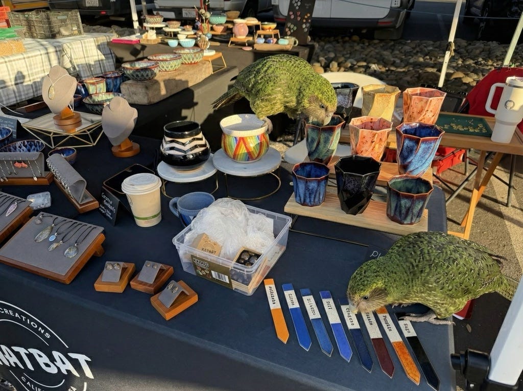 Same craft market booth with two Kākāpō now in different positions: one remains center-table peering into the ceramic cups near the rainbow pot, while the second has moved to the right edge of the table near the plant markers, appearing to examine or possibly chew on items at the table's corner. They are both a little smaller than in the first image.
