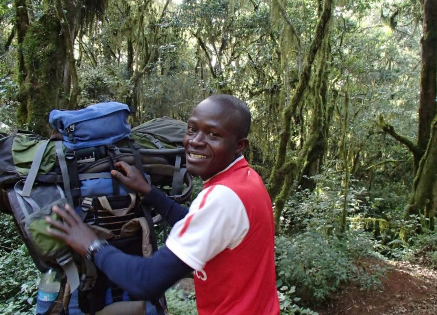 A young Tanzanian porter in a red and white soccer jersey smiles over his shoulder while carrying a large loaded backpack, surrounded by green moss-draped rainforest on the lower slopes of Kilimanjaro. A young Tanzanian porter in a red and white soccer jersey smiles over his shoulder while carrying a large loaded backpack, surrounded by green moss-draped rainforest on the lower slopes of Kilimanjaro.