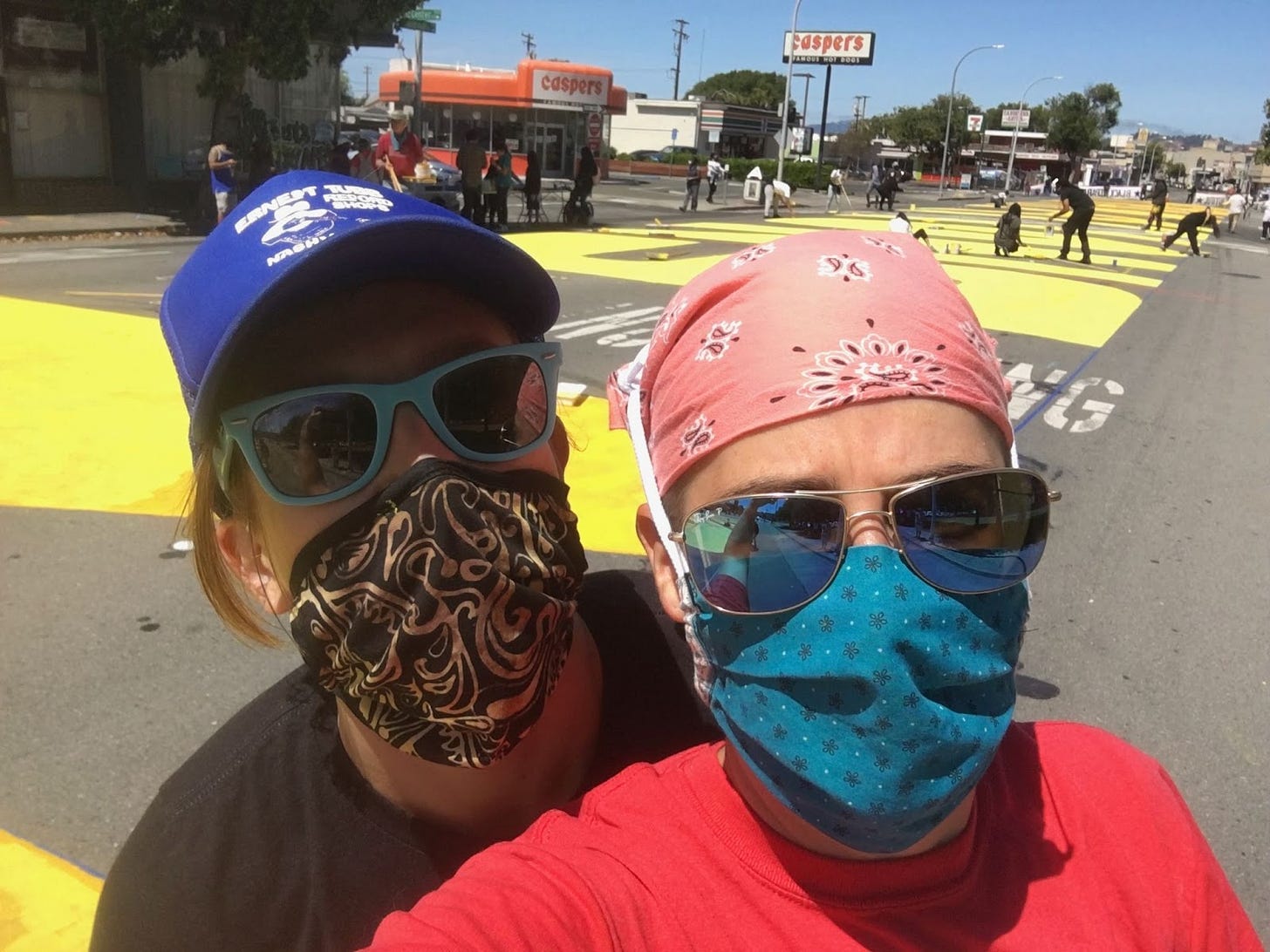 close up photo of my partner and I in the streets of the East Bay, posing in front of big yellow letters painted on the ground that read “Black Lives Matter”. We are wearing sunglasses, hats and bandanas on her heads, and Covid 19 masks.