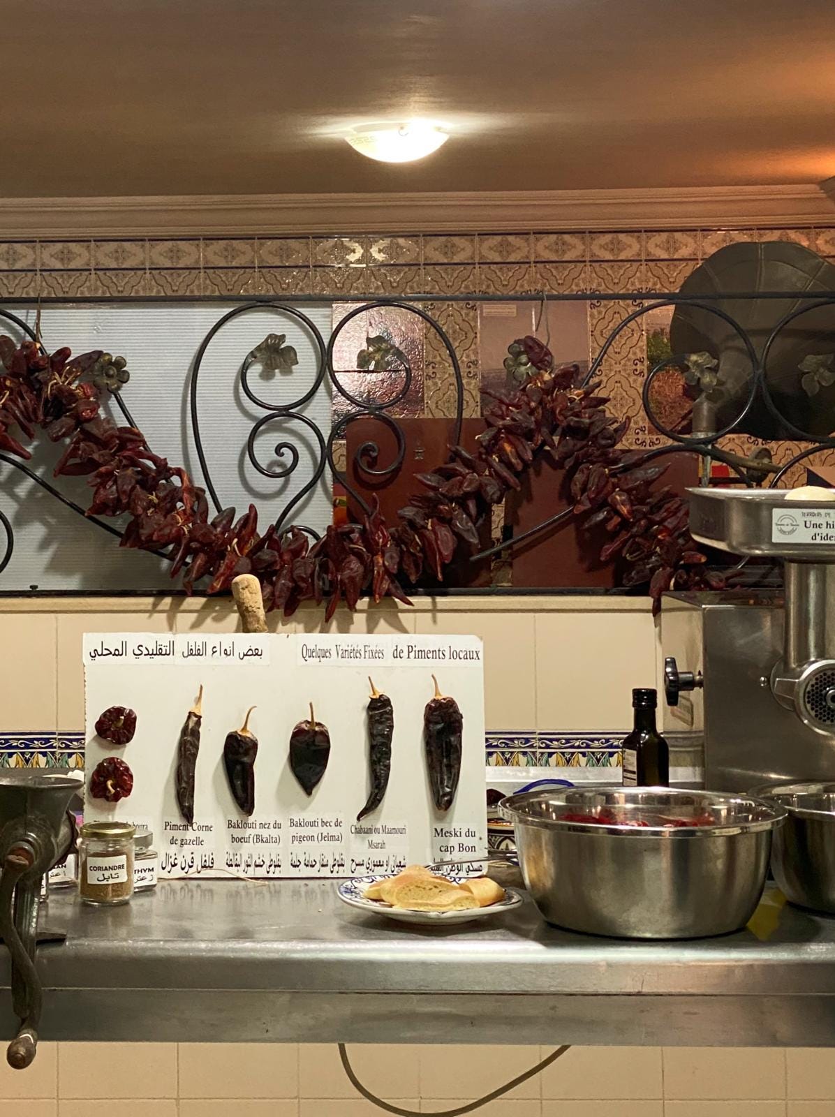 A chrome silver table in a basement with a garland of chillies behind, and a display board with dried chillies pinned to it, as well as the equipment for making harissa