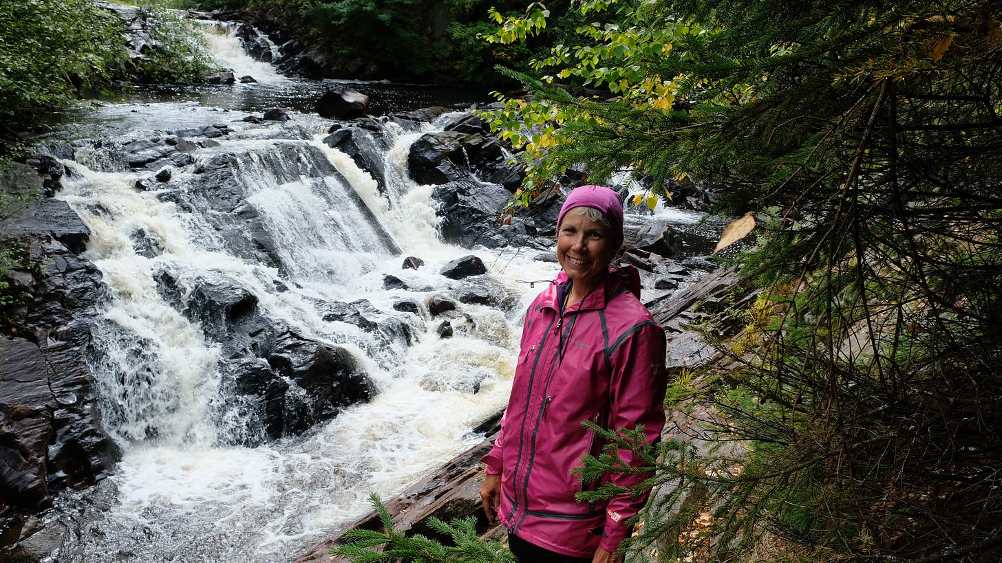 Vicki, dressed in hot pink waterproof cool weather gear, standing in front of a rushing fall of water in a stone-bedded creek. Lush green trees line the creek.