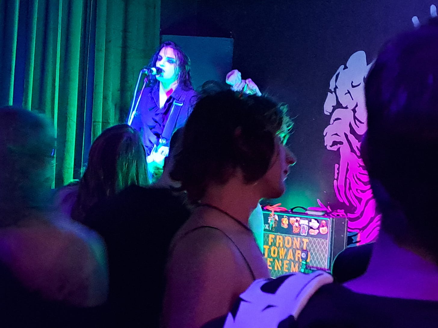 A man in goth attire and a ruffled shirt singing in front of a crowd in a darkened room.