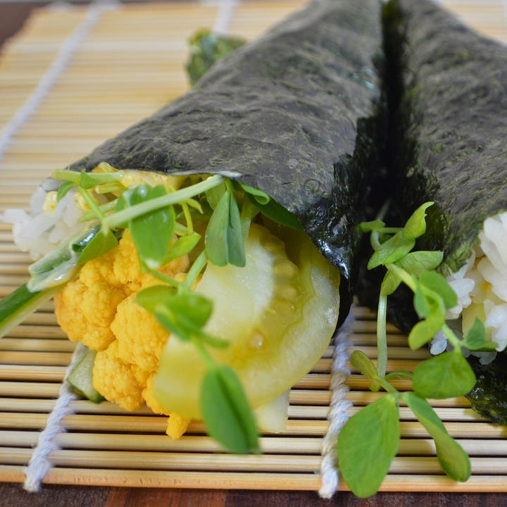 Two hand rolls (temaki): one shaped like a traditional cone with nori wrapped around rice and fillings, and the other served open-faced in a taco-style presentation. Both are plant-based and displayed side by side.