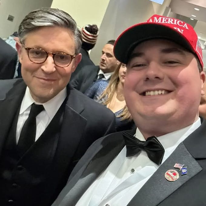 Two men pose smiling indoors at a formal event; the man on the left wears glasses a dark suit white shirt black bow tie and has gray hair; the man on the right wears a red Make America Great Again cap a white shirt with American flag and DNC pins and a black bow tie; a woman in a red dress and another man in a suit stand blurred in the background.