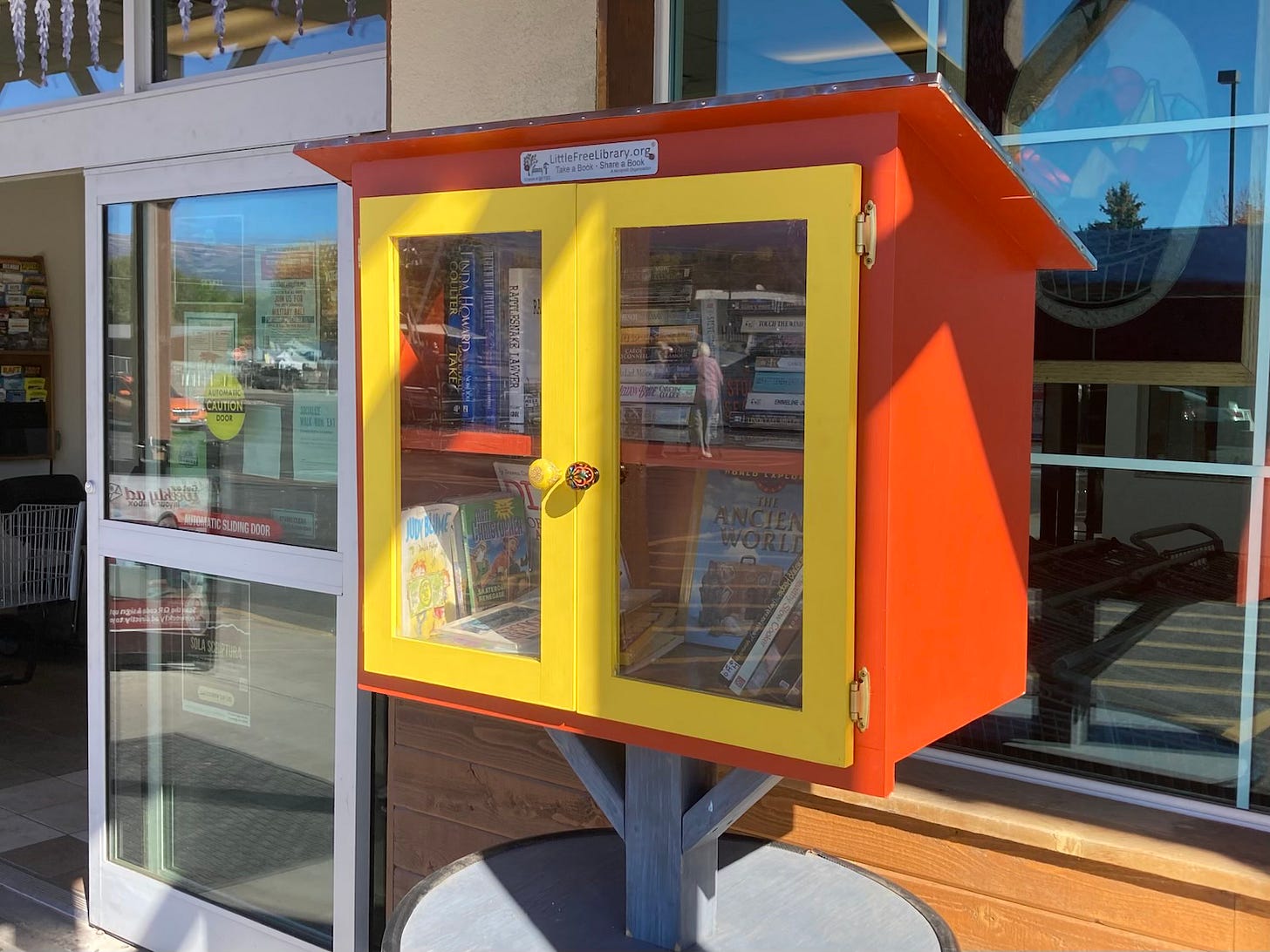 A red box on a post at the entrance to a grocery store. The front has two yellow doors with windows. Inside the box there are books. 