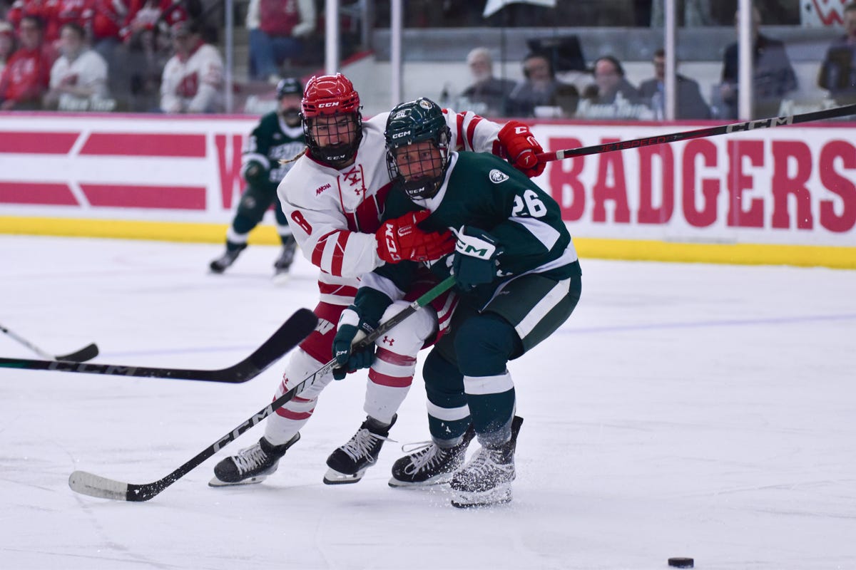 A hockey skater in a white Wisconsin Badgers women's hockey jersey attempts to skate past a Bemidji State hockey player in a green jersey as the BSU player turns their hips up ice A hockey skater in a white Wisconsin Badgers women's hockey jersey attempts to skate past a Bemidji State hockey player in a green jersey as the BSU player turns their hips up ice