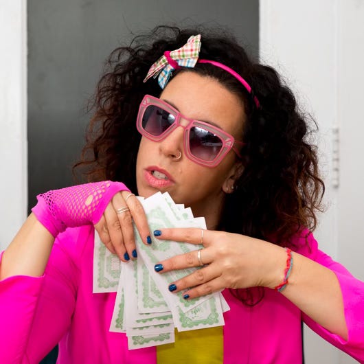 Woman in a hot pink blazer, pink sunglasses, and a bow headband holding a fanned stack of bills with a skeptical expression — because your money story is complicated, and so is hers.