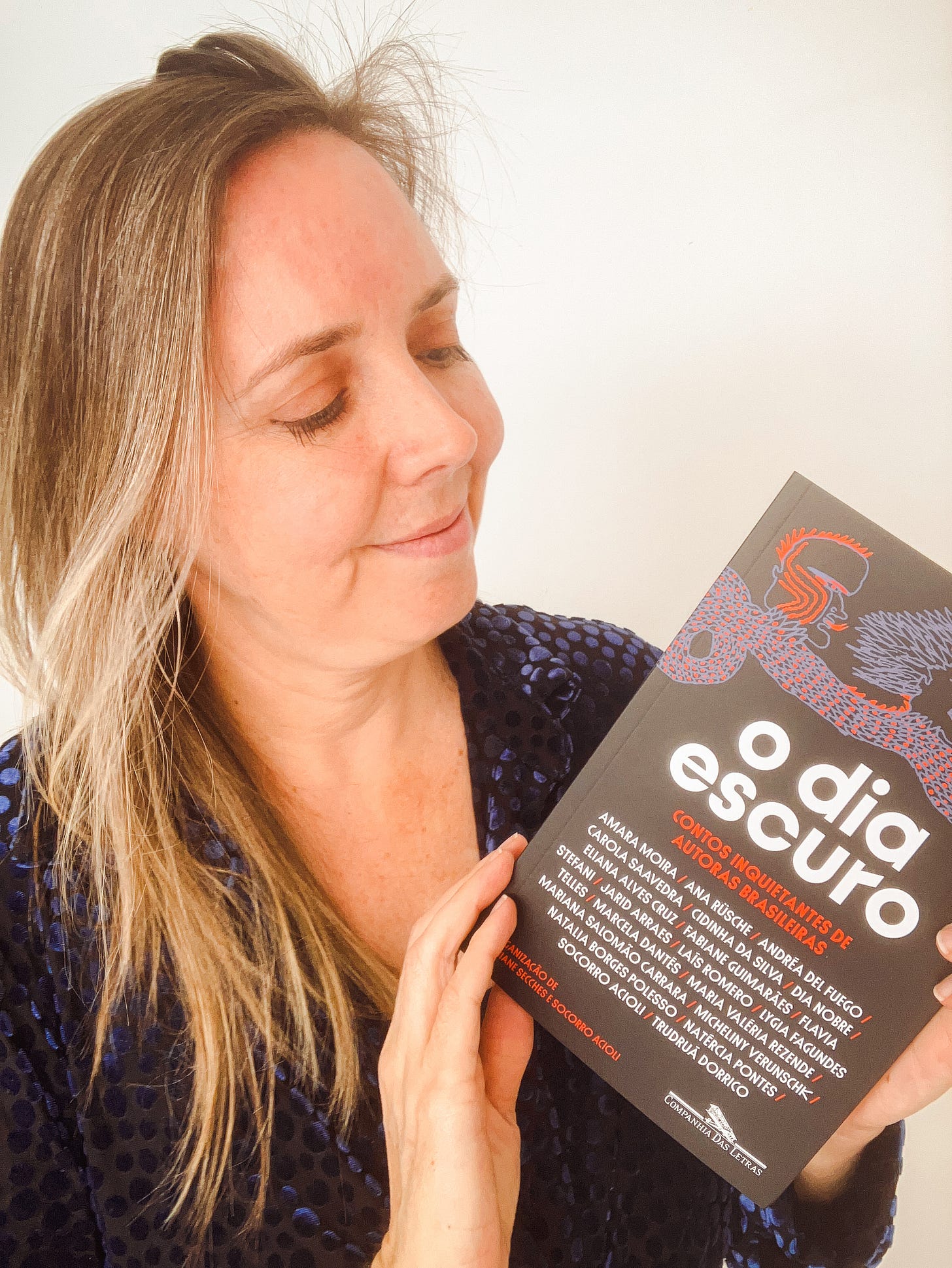 Uma mulher branca, com cabelos claros e lisos de meia idade, segura um livro de capa preta com desenhos de monstros lilases, com o tútulo "O dia escuro"