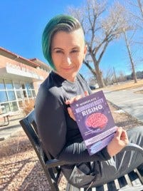 Lyric, a pale skinned multiracial, nonbinary human, with short green hair and shaved sides is sitting on a bench in front of a brick building that says "school of business" - in Lyric's hand is a purple book with a pink brain on it that says Workplace NeuroDiversity Rising Lyric, a pale skinned multiracial, nonbinary human, with short green hair and shaved sides is sitting on a bench in front of a brick building that says "school of business" - in Lyric's hand is a purple book with a pink brain on it that says Workplace NeuroDiversity Rising