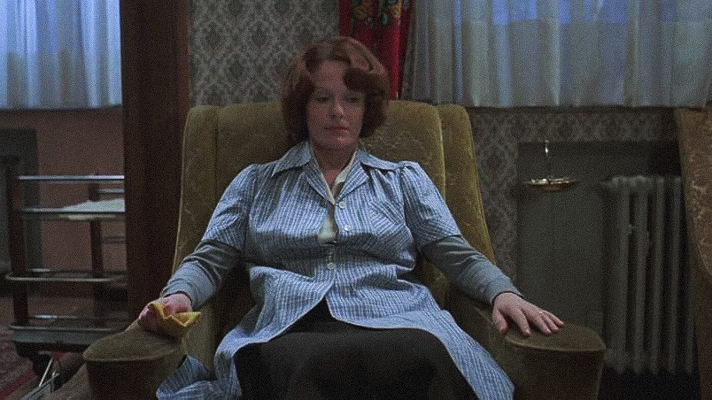 Jeanne Dielman sits motionless in an armchair, staring downward with a blank expression, framed by the muted tones of her modest living room.