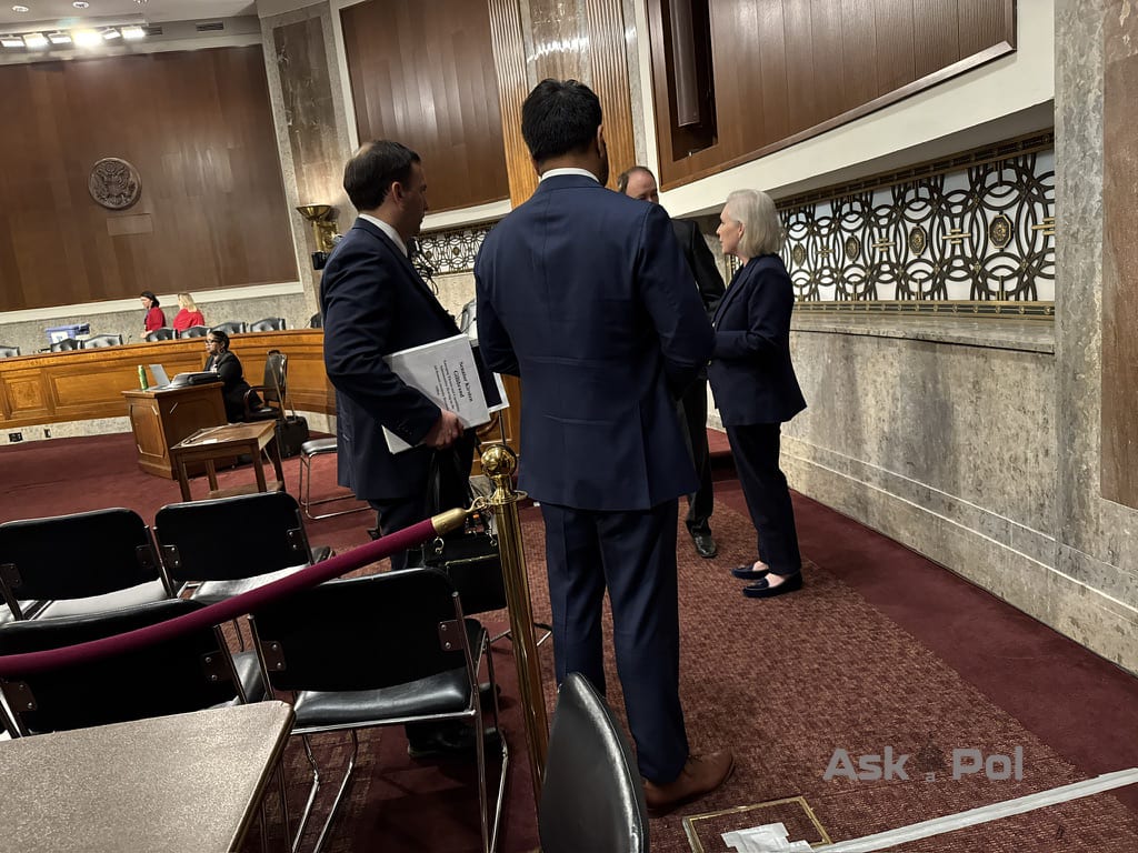 Men in suit stand around a female Senator in a large committee hearing room Photo: Matt Laslo © www.askapolcrypto.com