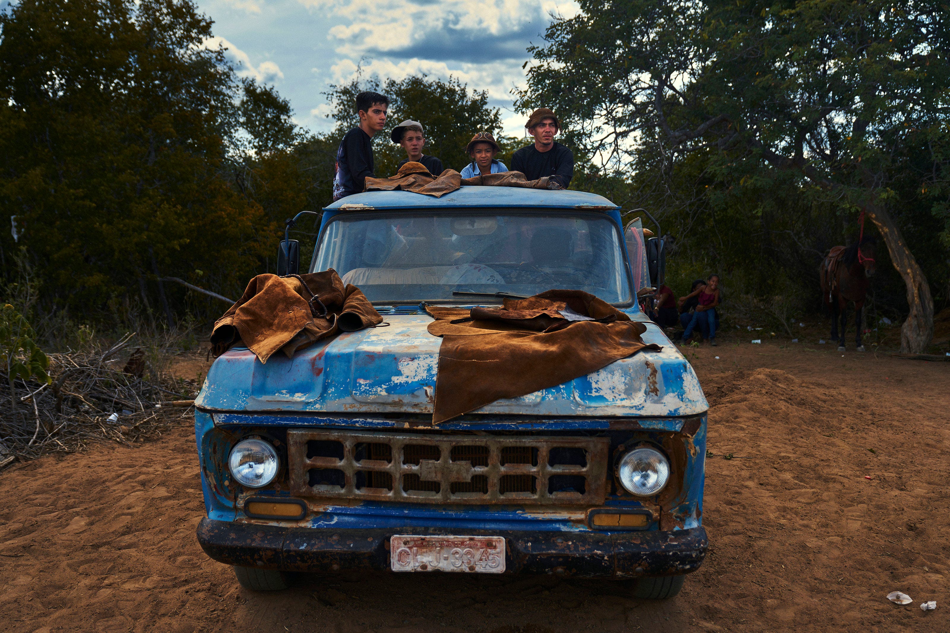 Four boys stand and sit on top of an old, weathered blue truck with brown blankets on the hood, surrounded by dirt and trees; two people and a horse are visible in the background.