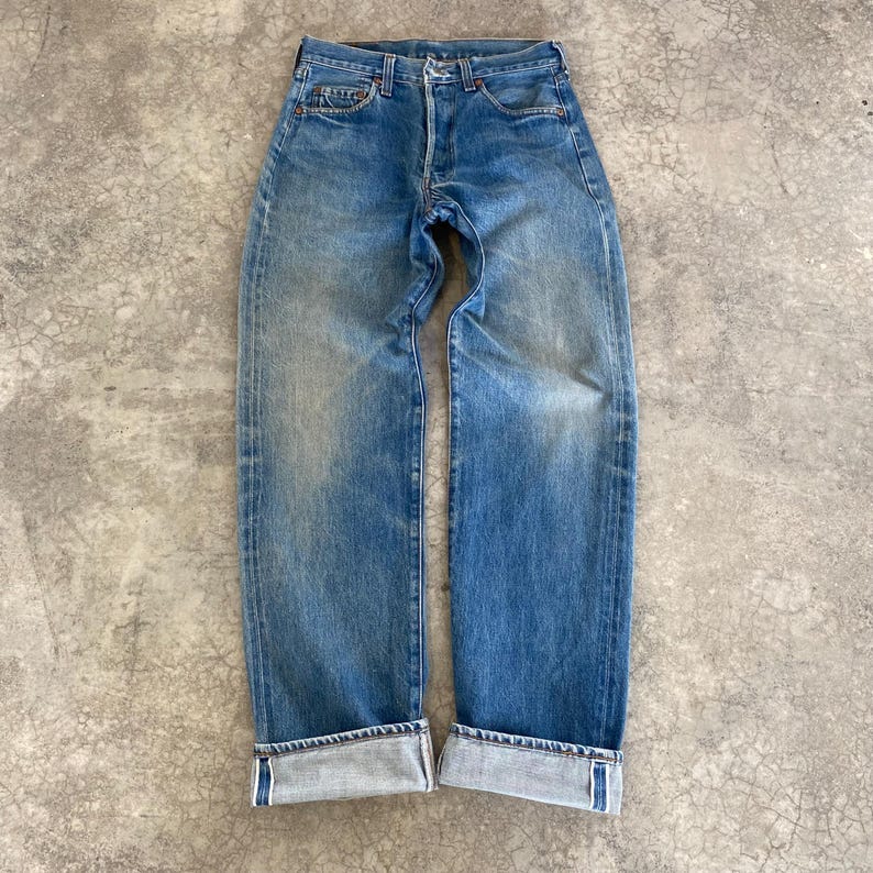 May include: Faded blue denim jeans with a button fly and straight leg. The jeans have a classic five-pocket design with visible stitching and a worn appearance. The cuffs are rolled up, revealing the interior fabric. The jeans are displayed on a gray, textured surface.