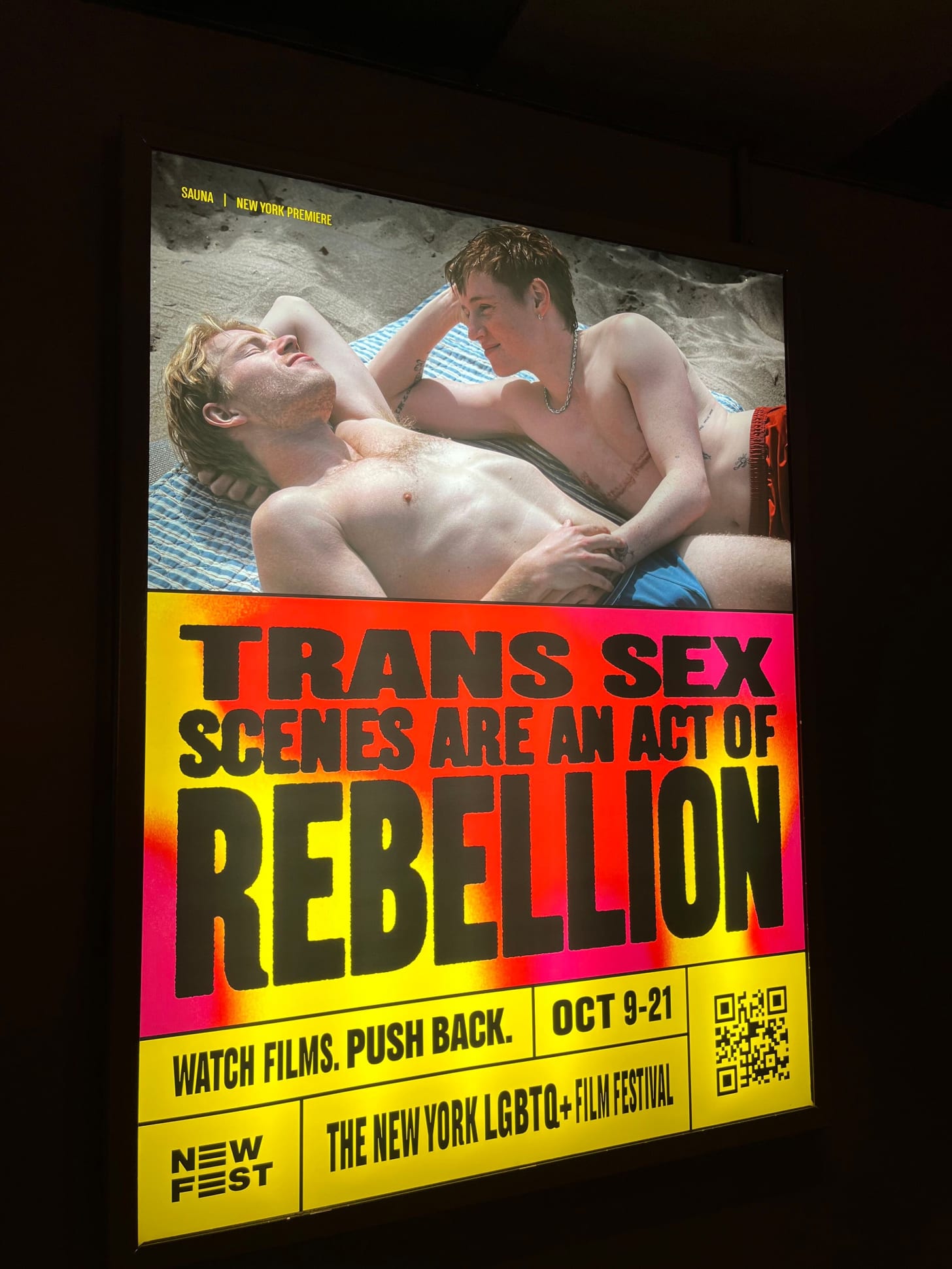 A back-lit sign shows a cis gay guy and a trans gay guy with top surgery scars lying on the beach. Underneath is a graphic that says TRANS SEX SCENES ARE AN ACT OF REBELLION. Underneath it says Watch Films. Push Back.