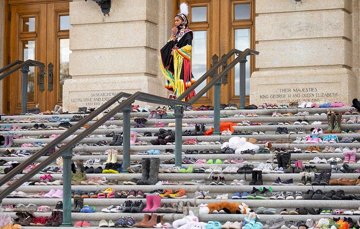 A person in colorful traditional dress stands on steps filled with shoes. Stone building in background. Text on stone mentions Saskatchewan. A person in colorful traditional dress stands on steps filled with shoes. Stone building in background. Text on stone mentions Saskatchewan.