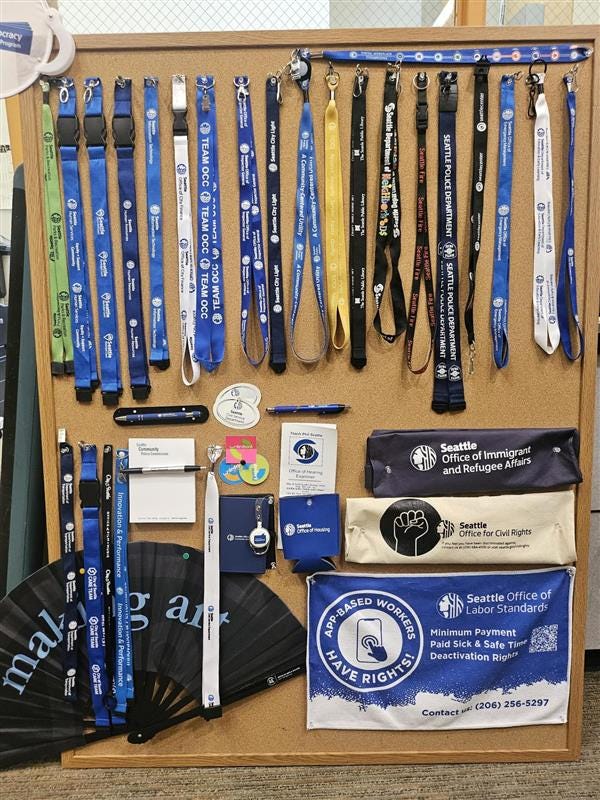 Corkboard with Lanyards, pens, stickers and other pieces of "swag" with printed logos of the City of Seattle with various department names on them. There are around 40 items. Corkboard with Lanyards, pens, stickers and other pieces of "swag" with printed logos of the City of Seattle with various department names on them. There are around 40 items.