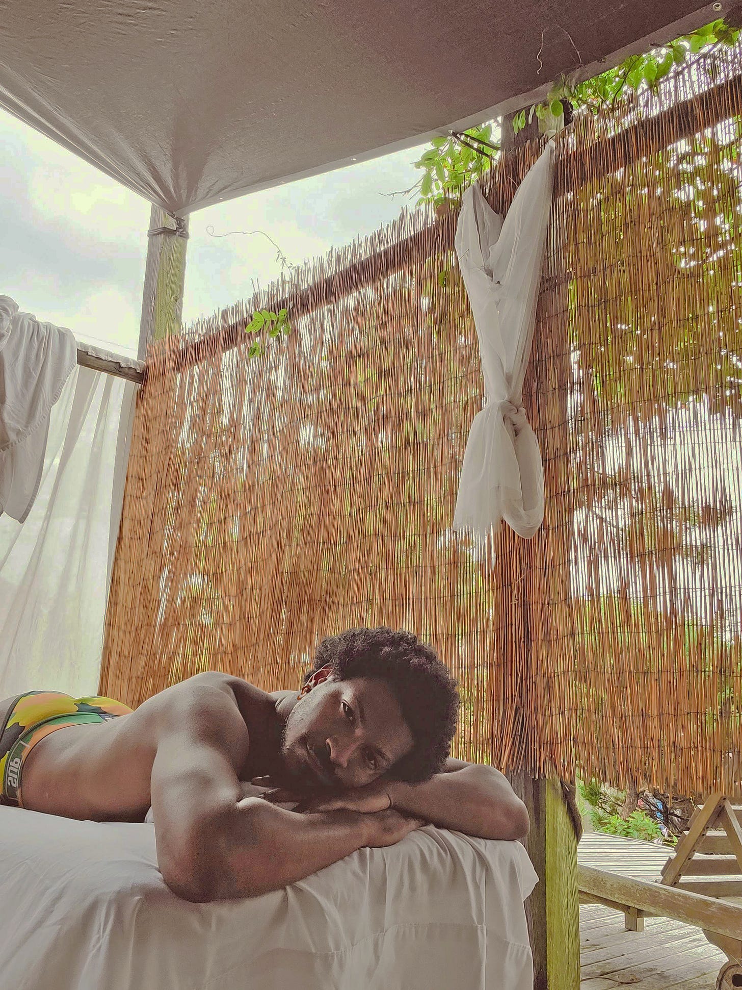 Person lying face-down on a massage table on an outdoor deck, head resting on folded arms, surrounded by bamboo screens and soft natural light. Person lying face-down on a massage table on an outdoor deck, head resting on folded arms, surrounded by bamboo screens and soft natural light.