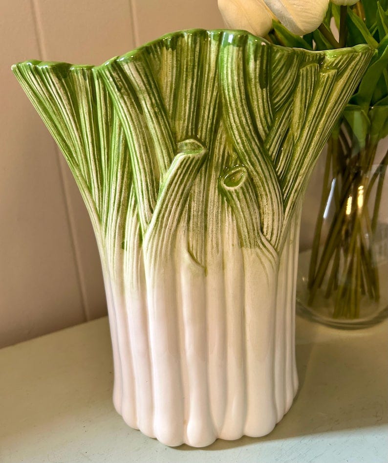 May include: A ceramic vase shaped like a leek with a white base and green top. The vase has a textured surface that resembles the leaves of a leek.