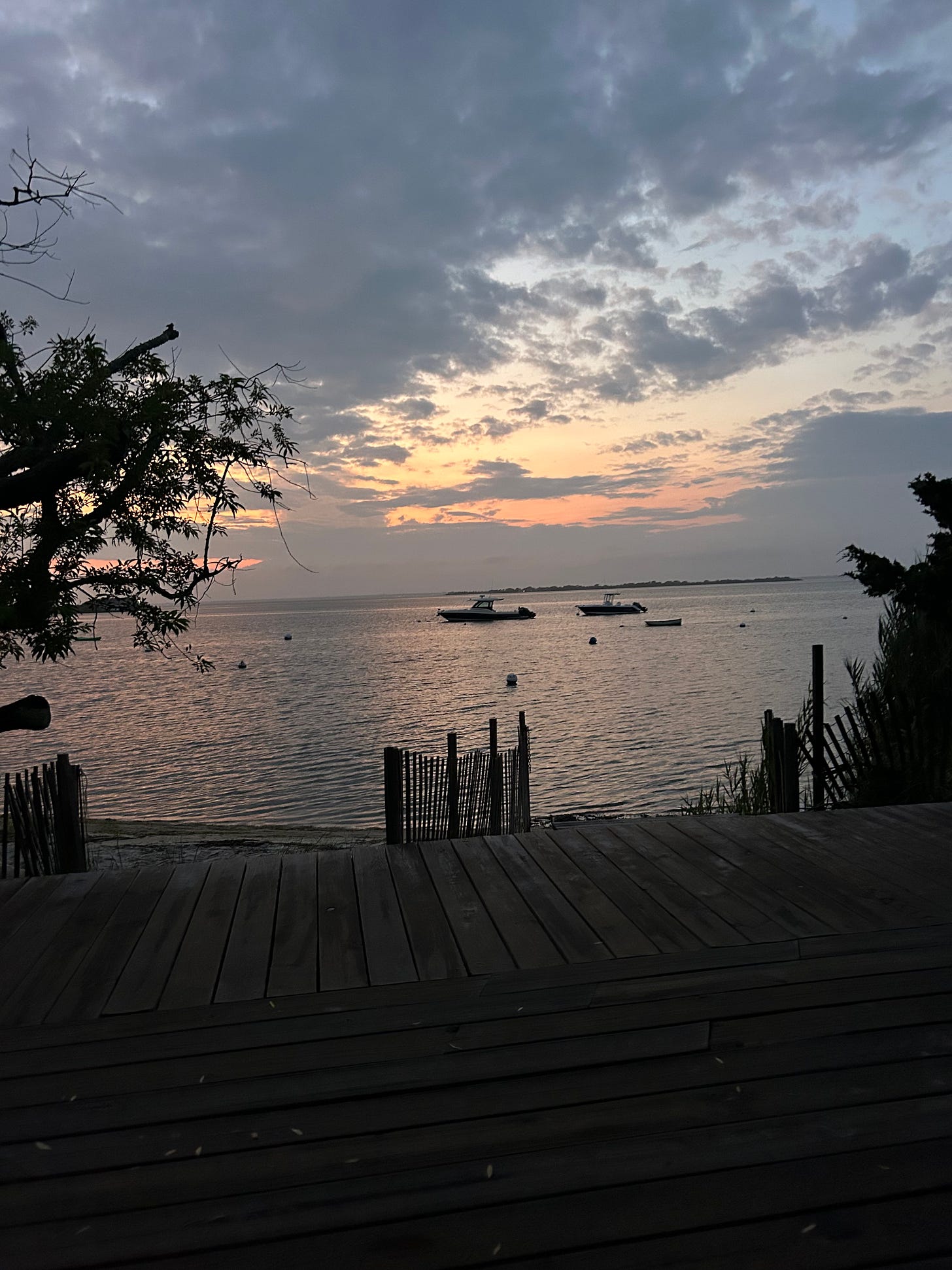 The view of the bay from the boardwalk in Fire Island at sunset. There are some small boats and a rickety wooden fence