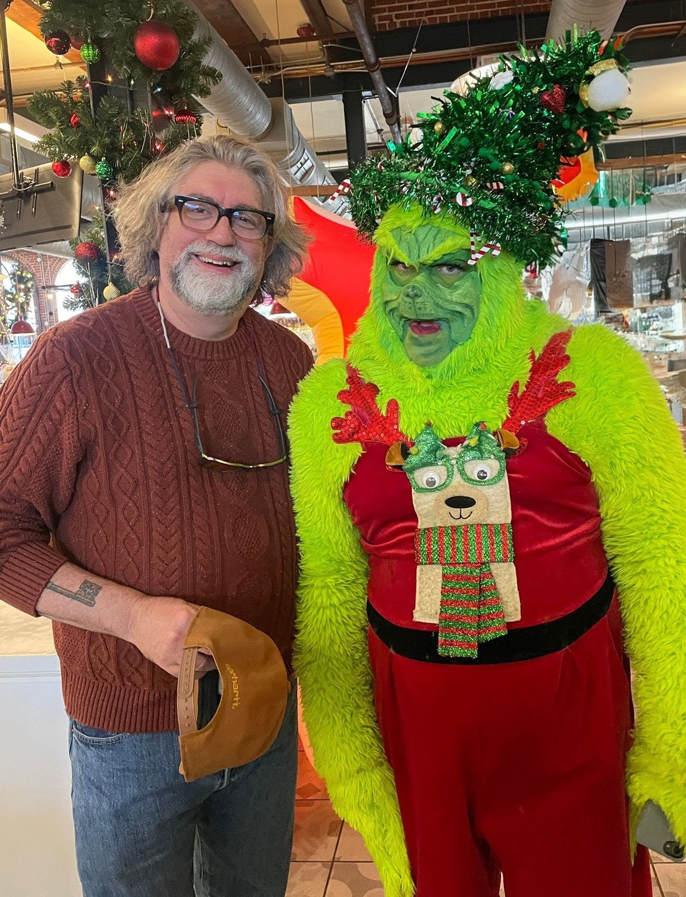 white man with salt and pepper beard and black glasses next to a man in a Grinch costume at a holiday event