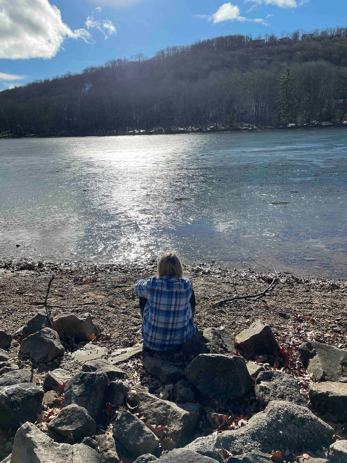 Cassidy in blue plaid facing a thinly ice covered lake. Leafless trees and houses are visible on the other side of the lake with a mostly blue sky. Cassidy in blue plaid facing a thinly ice covered lake. Leafless trees and houses are visible on the other side of the lake with a mostly blue sky.
