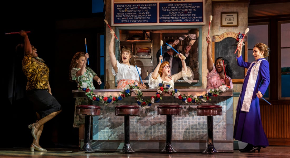 A stage with a group of people stood by a diner counter, holding spoons up A stage with a group of people stood by a diner counter, holding spoons up