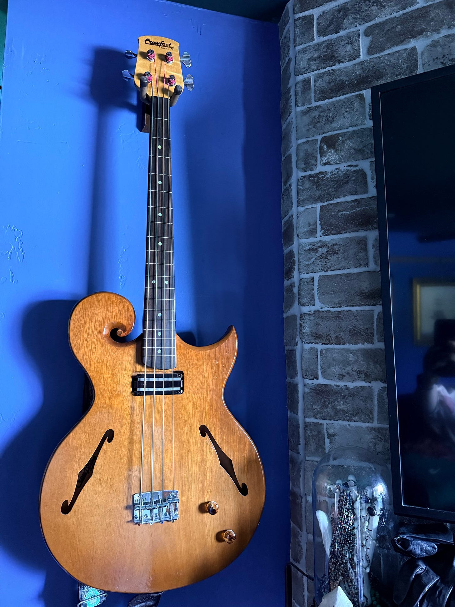 A natural wood four-string electric bass guitar with a distinctive curled body and F-shaped sound holes hangs against a vivid blue wall beside exposed grey brickwork. A natural wood four-string electric bass guitar with a distinctive curled body and F-shaped sound holes hangs against a vivid blue wall beside exposed grey brickwork.