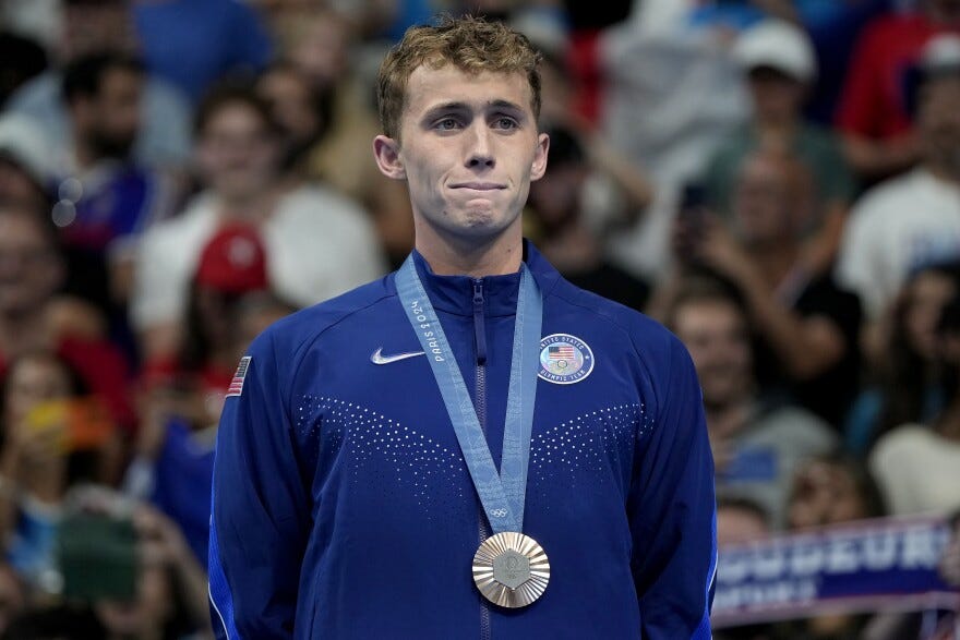 a man stands with his hands behind his back wearing a blue jacket and a ribbon medal around his neck