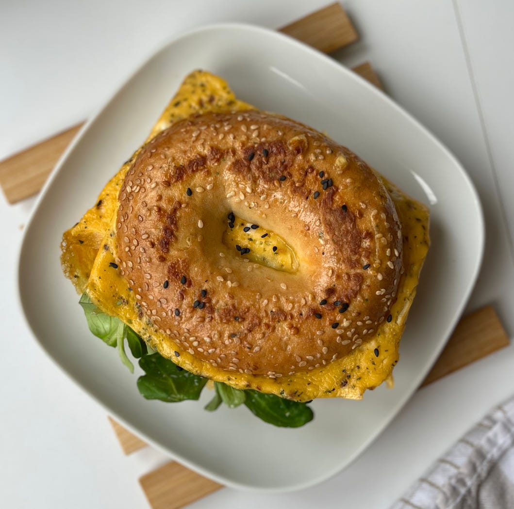 A delicious breakfast bagel sandwich filled with fluffy eggs, creamy cottage cheese, fresh avocado slices, and lamb’s lettuce, served on a white plate.