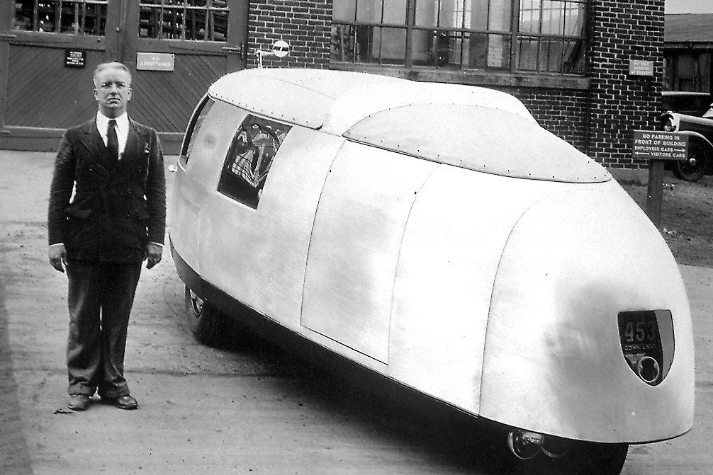A man stands next to an ovoid car with a smooth exterior. 