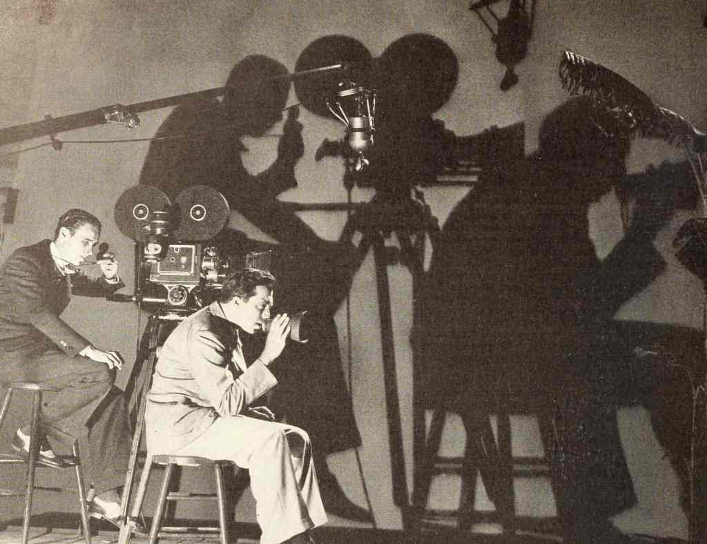 Black and white photograph of two men with 1930s film camera casting large shadows on the wall. One man is speaking into a loudspeaker