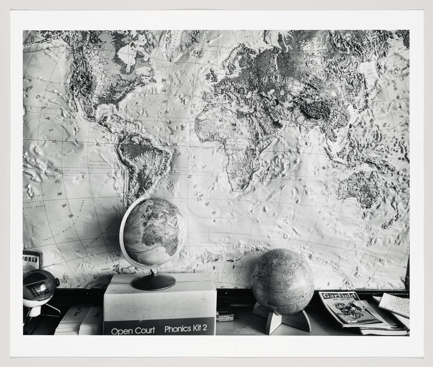 A black and white photo of a study area with a large world map on the wall, two globes on a shelf in front of the map, and various educational materials including books and a phonics kit.