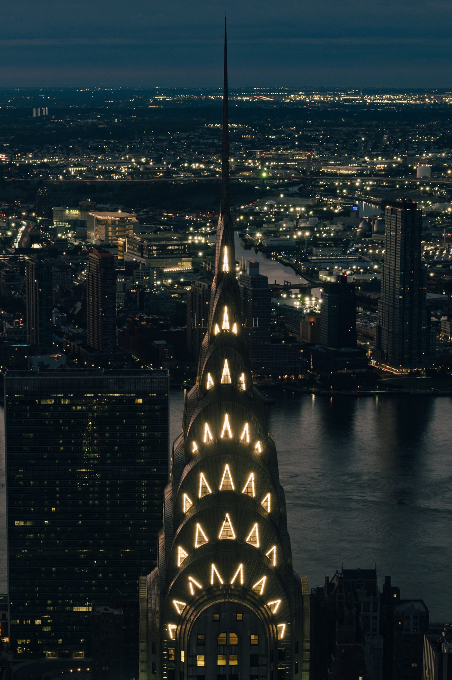 The Chrysler Building lit gold at night seen from above, the dark East River and the lights of Queens behind it. The Chrysler Building lit gold at night seen from above, the dark East River and the lights of Queens behind it.