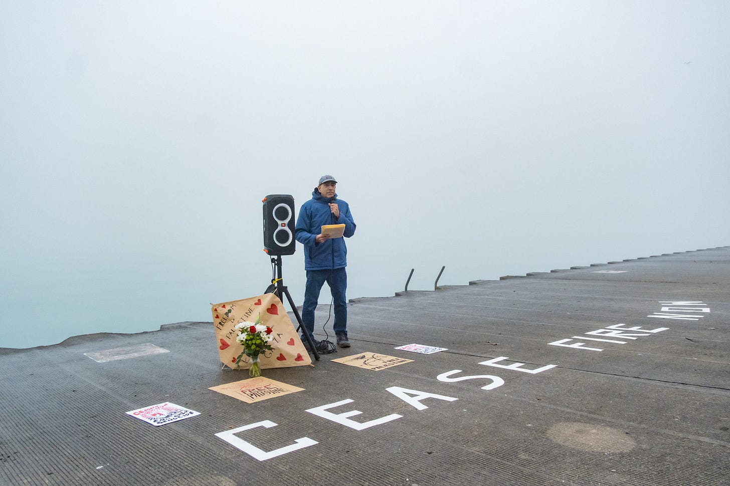 A man speaks into a microphone in front of a lake. Beside him is a speaker and a kite with the words Free Palestine written on it.