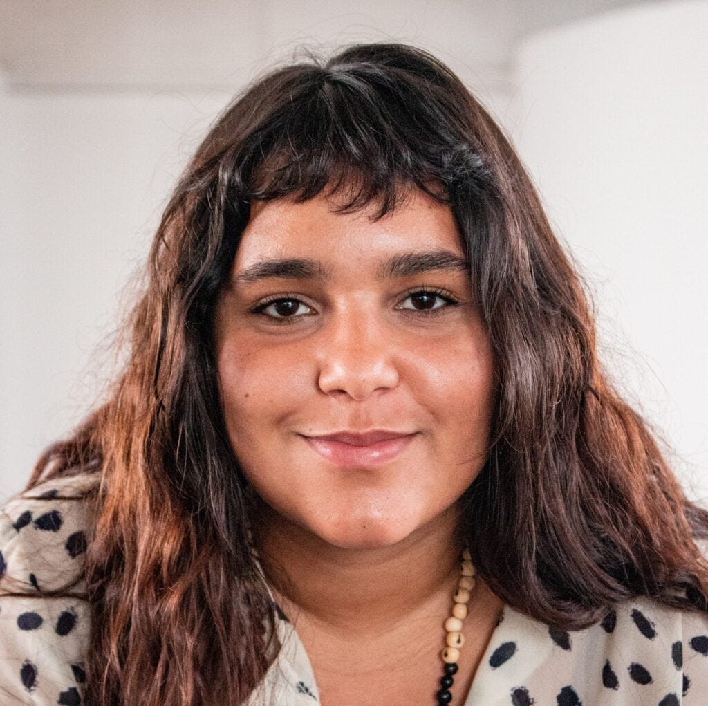Foto de Gabriella, do Prato Firmeza, mulher indígena com cabelo castanho longo e franja curta, vestindo uma camisa branca com bolinhas pretas.