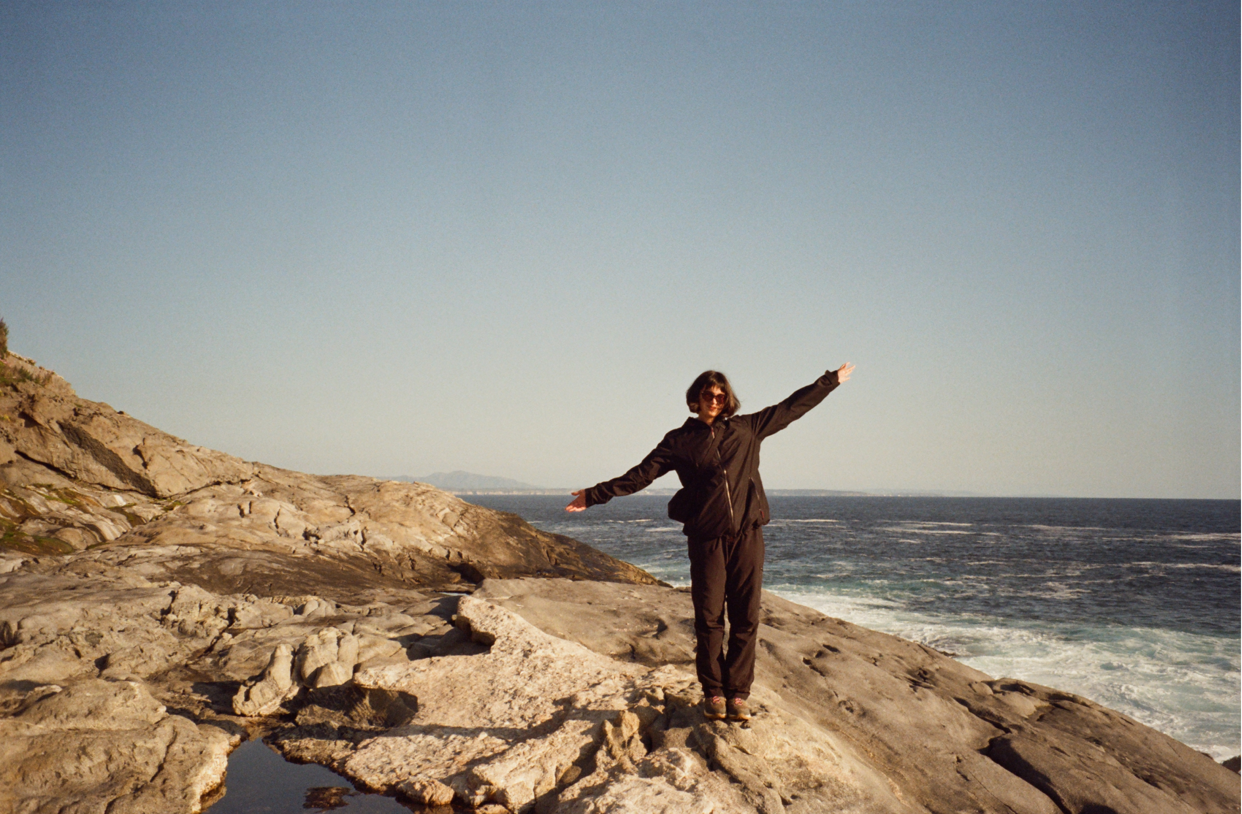Laura Sandoval stands with her arms in a tilted T on a rock in front of the ocean.