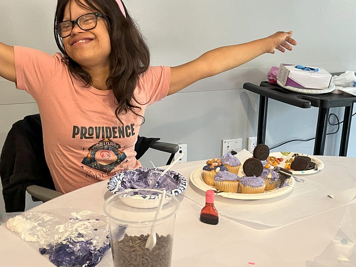 James is a middle-aged man who holds a plate of decorated cupcakes. Taylor is a young woman with long hair and glasses who sits beside her plate of decorated cupcakes.