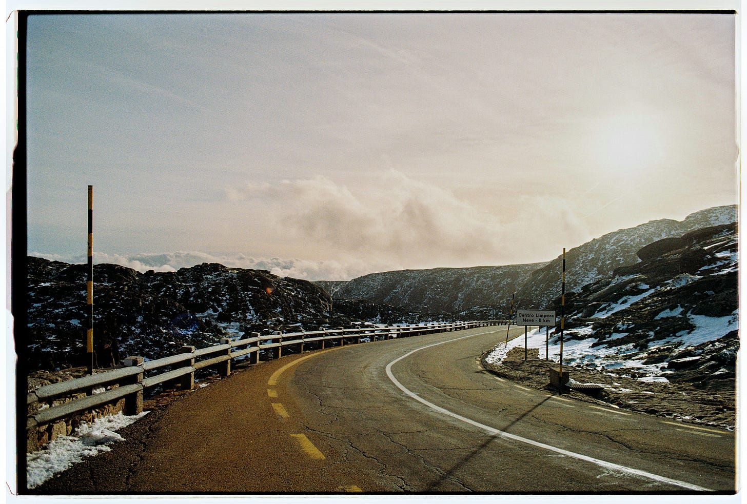 Mountain road curves through a rugged landscape with guardrails guiding the path ahead under a bright sky.