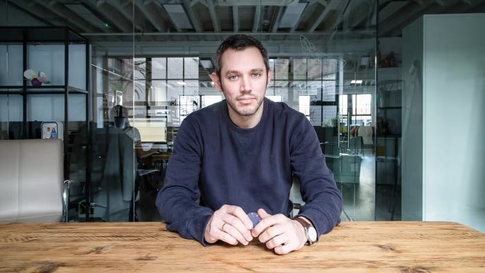 Jake is sitting at a wood table in an office space looking directly at the camera. He has short grey and black hair. He is wearing a blue sweatshirt. He has white skin. He has a short beard. His hands are on the table. He is leaning forward slightly. He is not smiling. 