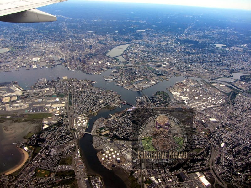 This is an aerial photograph taken from an airplane, with the tip of a wing visible in the top left corner. The image shows a sprawling metropolitan area, with a large river or bay system cutting through it. There are numerous buildings, residential areas, highways, and industrial sites visible, along with several bridges spanning the water. The urban landscape gives way to more suburban and green areas in the distance. The photo appears to be of Boston, with its distinctive waterways and urban layout.