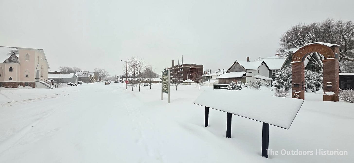 A view of the Italian Hall memorial site from the side, looking south down a quiet, snow-covered 7th Street in Calumet. The lot is empty and white, reflecting a sense of modern stillness.
