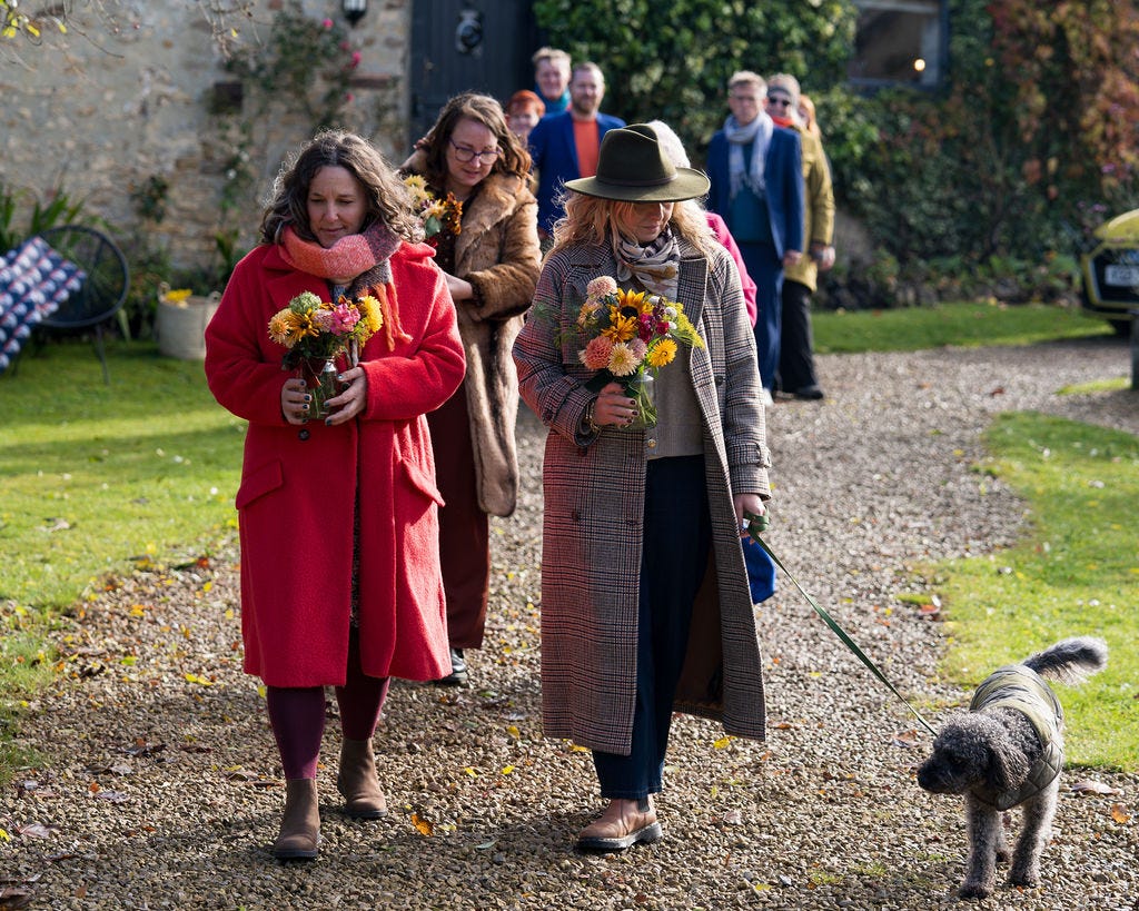 A procession of mourners and a dog walking down a garden path. The people are carrying jam jars of flowers cut from the garden