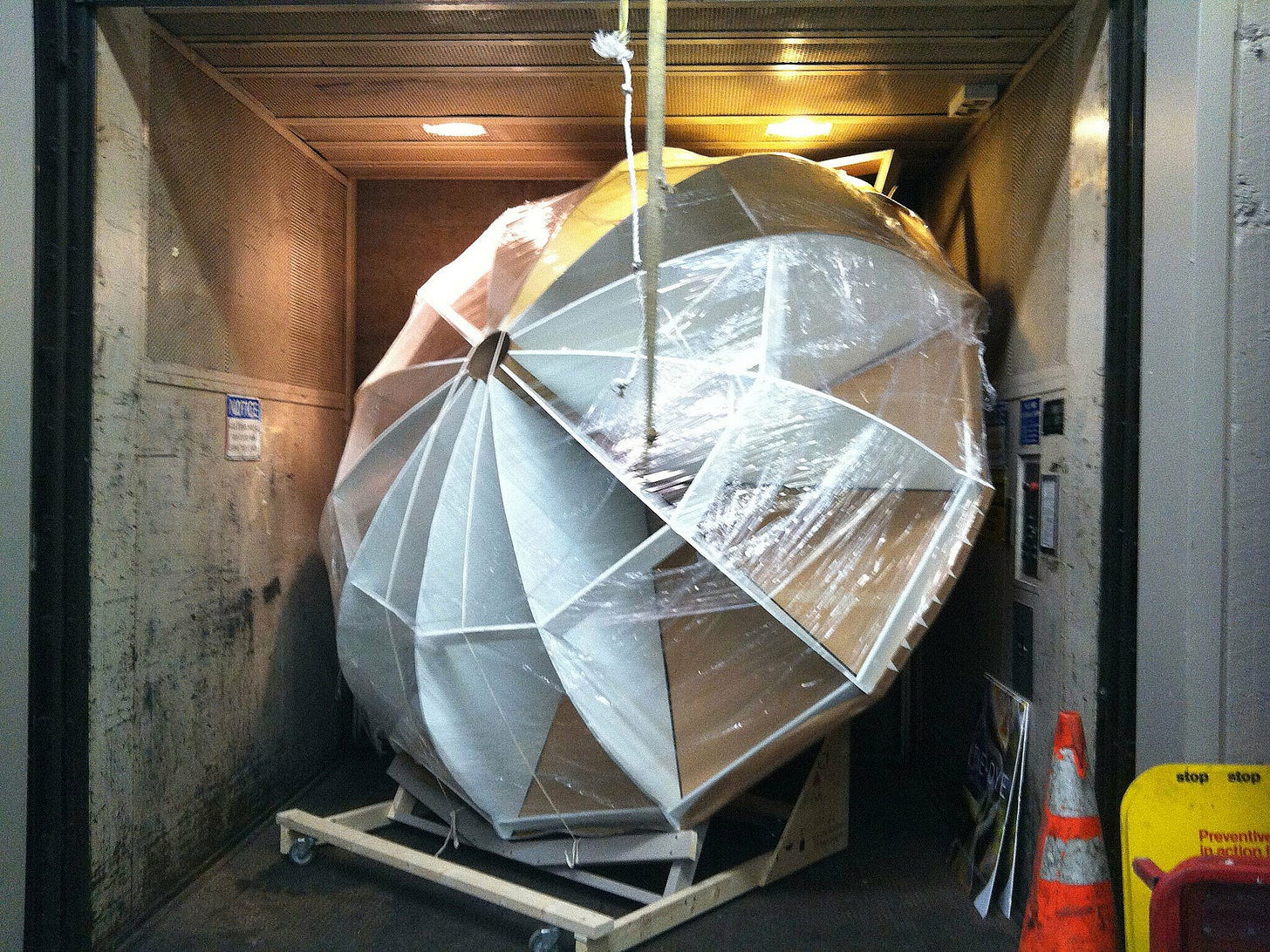 Large geometric structure wrapped in plastic, tightly packed inside an elevator. Orange safety cones and signs are visible nearby.